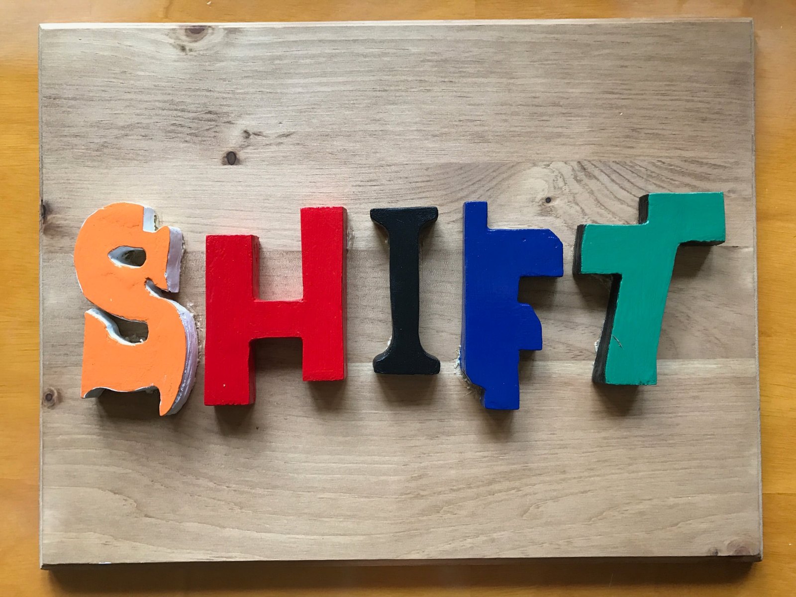 Handmade wooden SHIFT letters painted orange, red, black, blue and green, mounted on a pine board