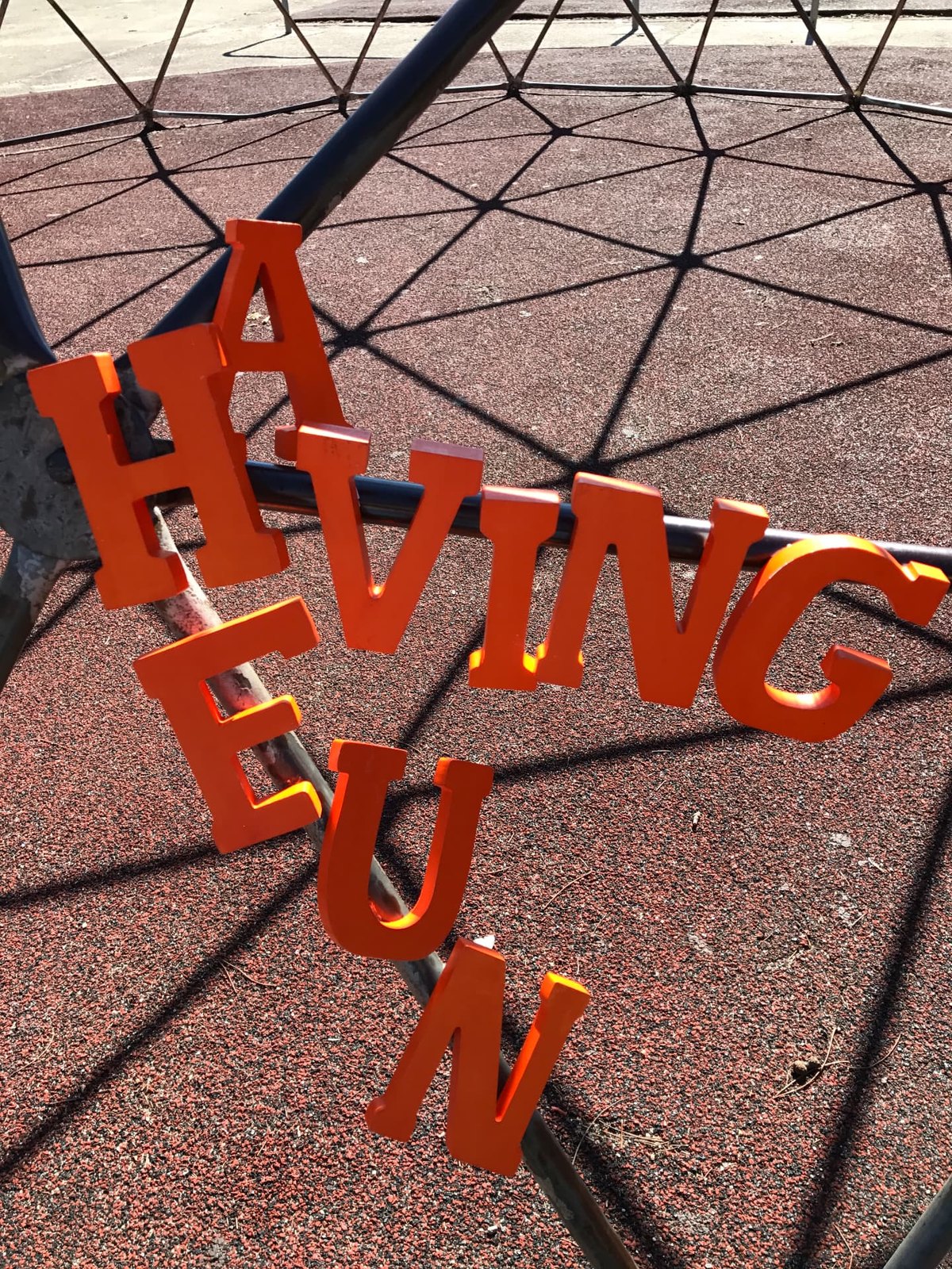 Orange wooden letters spelling HAVING FUN hung along the bars of a playground climbing dome