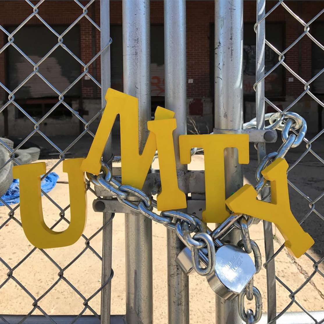 Yellow wooden letters spelling UNITY hung across a chained and padlocked metal gate