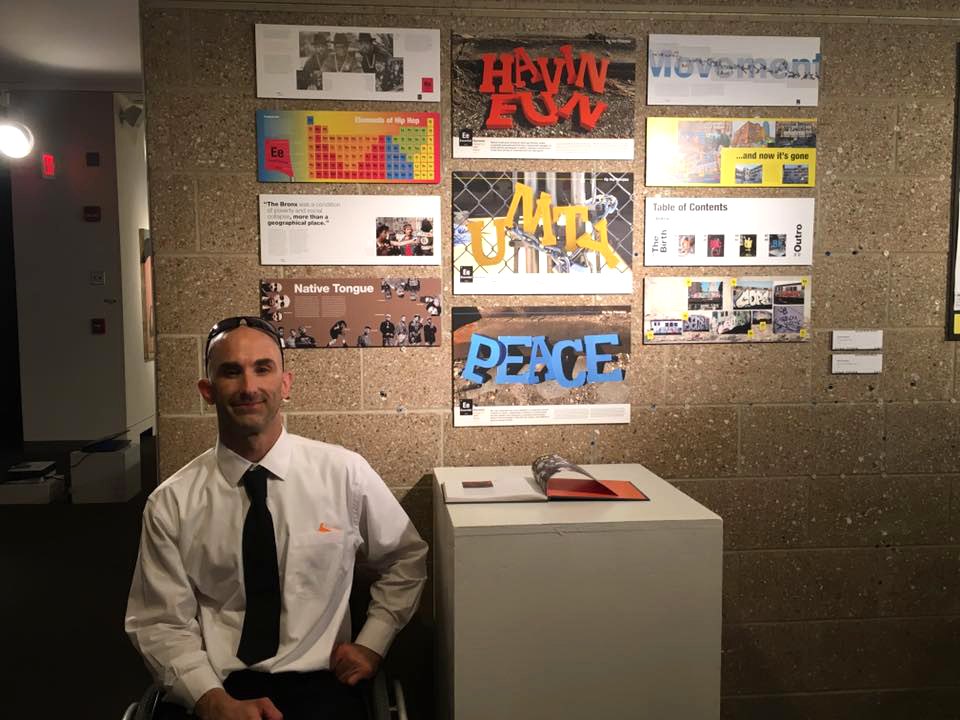 David Gardner seated in front of a wall of his exhibited graphic design panels, with his printed book on a pedestal beside him