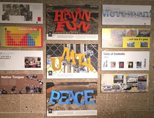Exhibition wall of Hip-Hop Principles panels, including an Elements of Hip Hop periodic table, a table of contents and photographs of the HAVIN FUN, UNITY and PEACE lettering