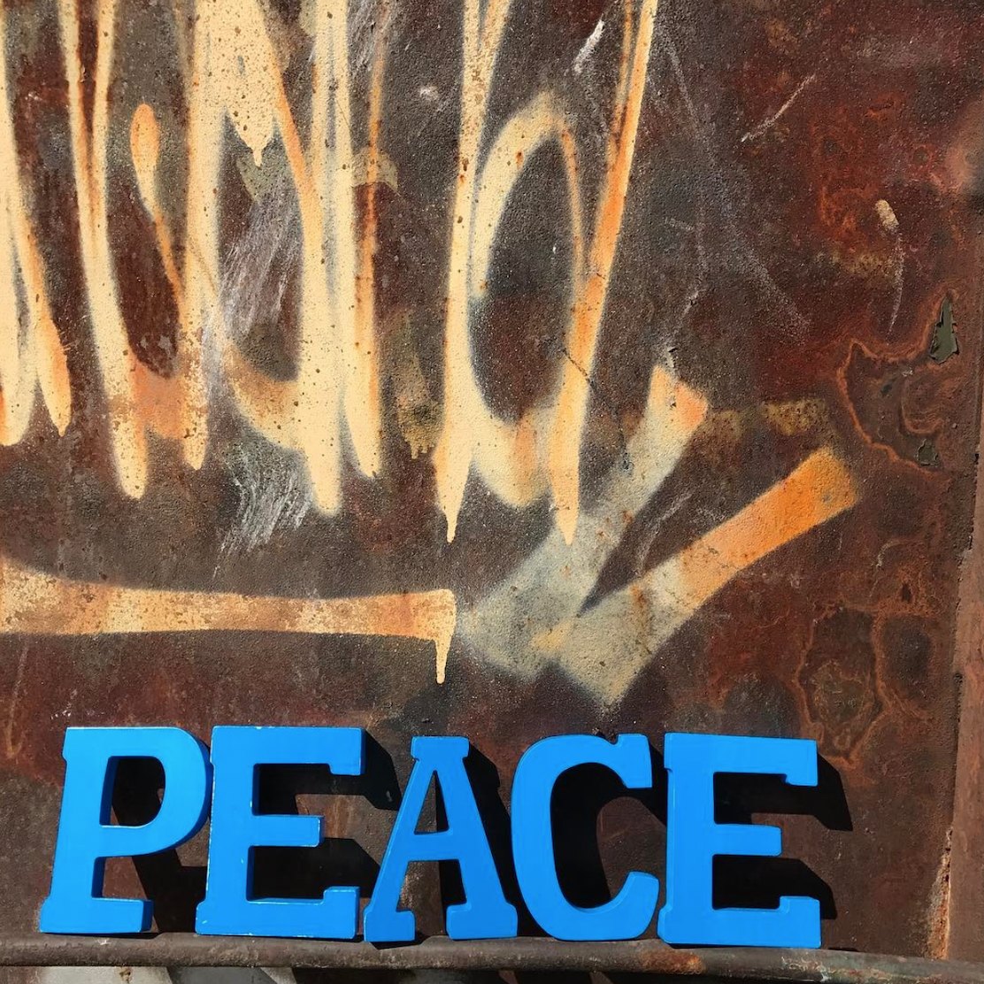 Blue wooden letters spelling PEACE set along the bottom of a rusted wall covered in spray paint