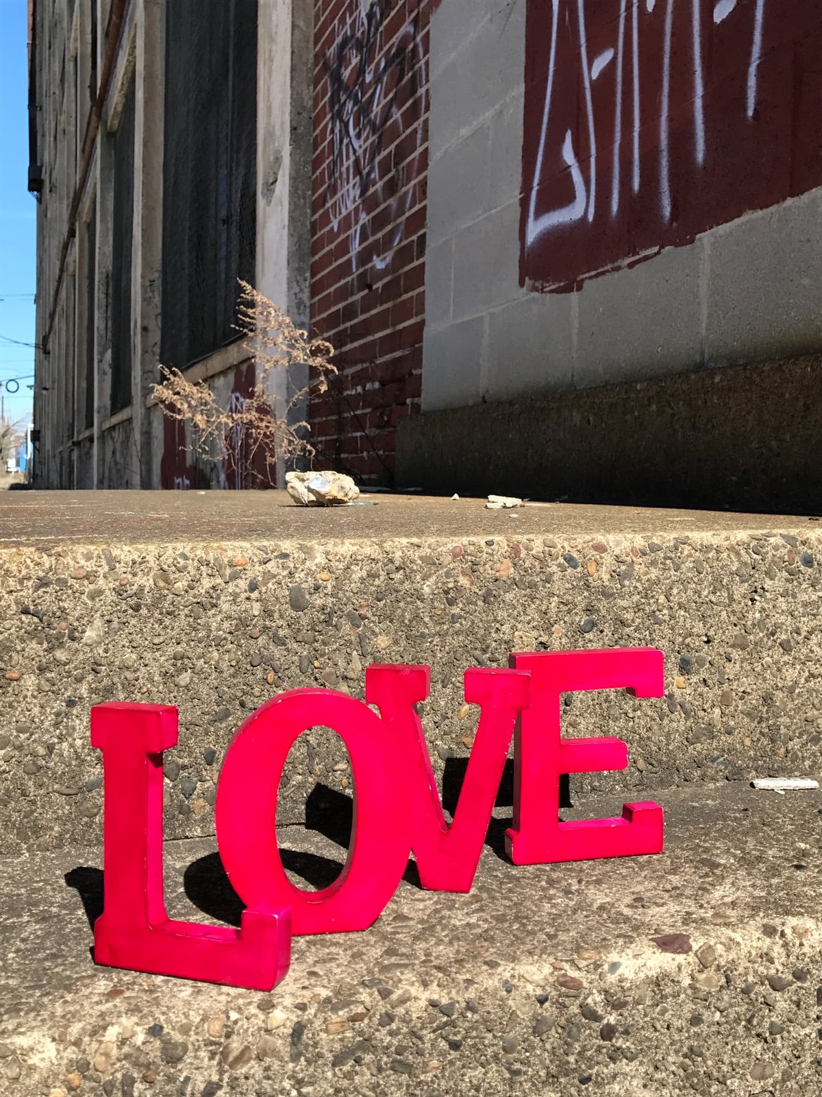 Bright pink wooden letters spelling LOVE propped on weathered concrete steps beside a graffiti-covered brick building